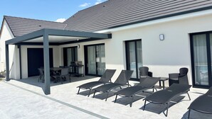 A covered outdoor seating area with lounge chairs, a table, and a grill.