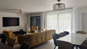 A modern living room with a sofa, chairs, a dining table, and a TV mounted on the wall.