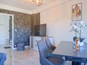 A modern living room with a flat-screen TV, a dining table with chairs, a small sofa, a decorative wall piece, and a potted orchid. - L'Auteuil by Interhome (Cagnes-sur-Mer)
