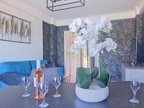 Uma sala de estar moderna com um sofá azul, uma mesa com copos de vinho e uma garrafa, e uma orquídea em vaso.