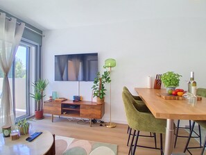 A modern living room with a flat-screen TV, a wooden dining table, green chairs, a floor lamp, and a variety of plants. - Le Tiercé by Interhome (Cagnes-sur-Mer)
