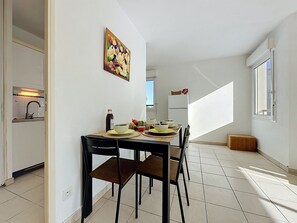 Ein Essbereich mit einem Tisch für vier Personen, eine Küche im Hintergrund und ein großes Fenster, das natürliches Licht hereinlässt.