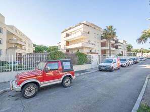 Ein rotes Auto steht auf einer Straße, im Hintergrund eine Reihe geparkter Autos und Wohnhäuser.