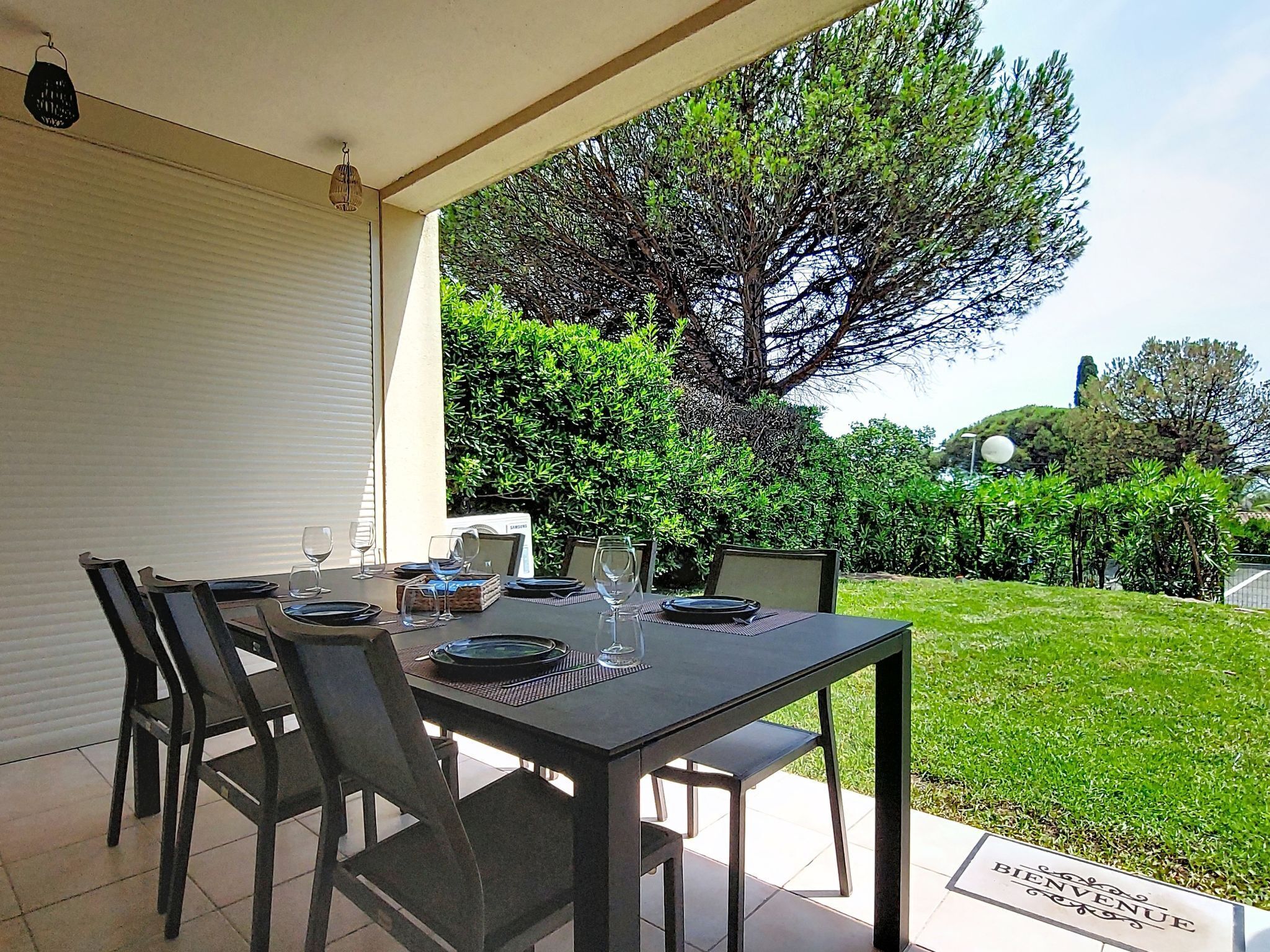 An outdoor patio with a table set for dining, surrounded by chairs, and a 'BIENVENUE' sign on the ground.