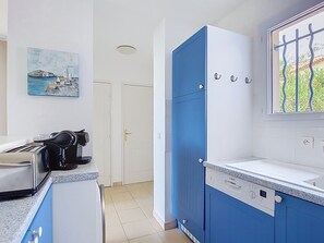 A kitchen with blue cabinets, a Bosch dishwasher, a window with a view of greenery, and a painting of a coastal scene. - Les Grenadines by Interhome (Les Issambres)
