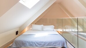 Ein Schlafzimmer mit schräg versiegelter Decke, einem Bett mit weißer Bettwäsche und einer Glasabtrennung.