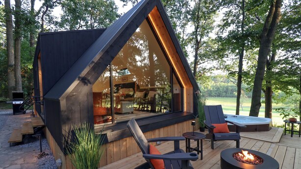 Een modern vakantiehuisje met een groot raam, houten terras, buitenmeubilair en een vuurkorf.