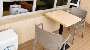 Un balcón con mesa y sillas, una cama con ropa de cama blanca, y vistas de la ciudad a través de amplias ventanas.