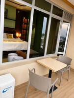 Un balcón con mesa y sillas, una cama con ropa de cama blanca, y vistas de la ciudad a través de amplias ventanas.
