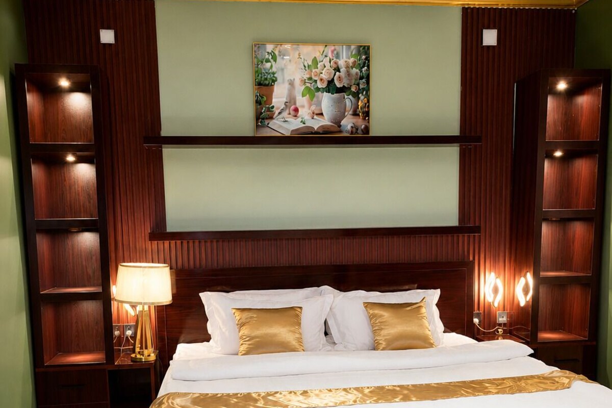 A large bed with a white and gold bedspread, flanked by wooden headboards and shelves, in a room with green walls and a ceiling with recessed lighting.
