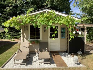 Ein kleines Haus mit überdachter Veranda, zwei Stühlen, einem kleinen Garten mit einer Vogelstatue und einem Schuppen mit einer Schwan-Dekoration.