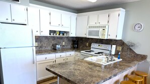 A kitchen with white cabinets, a microwave, a stove, a sink, and wooden stools. - Posh Piper: 2BD, 2BA Oceanfront Condo in Royal Garden Resort, Garden City (Murrells Inlet)