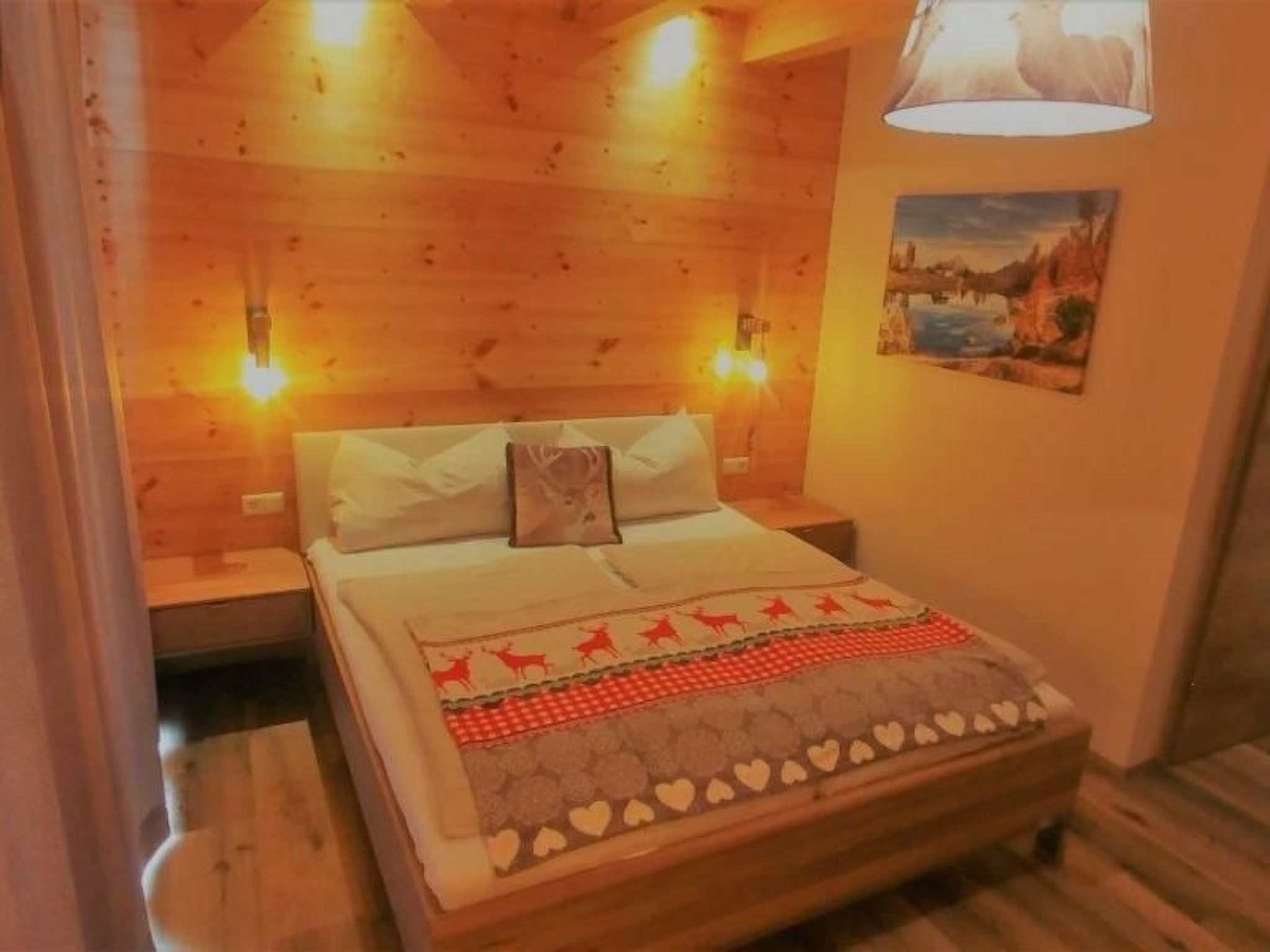A cozy room with a wooden ceiling, a bed with a patterned bedspread, two wall-mounted lamps, and a painting on the wall.
