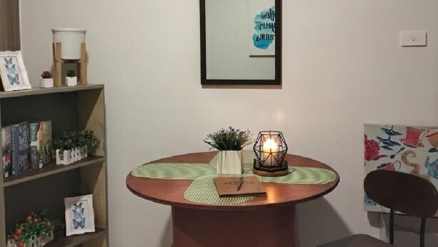 Una mesa redonda con una vela, una pequeña planta y una libreta. Se ve una estantería con libros y objetos decorativos.