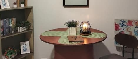 Una mesa redonda con una vela, una pequeña planta y una libreta. Se ve una estantería con libros y objetos decorativos.