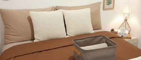 Un lit bien fait, recouvert d’une couverture marron, avec deux oreillers beige et une boîte de rangement grise posée sur le lit.