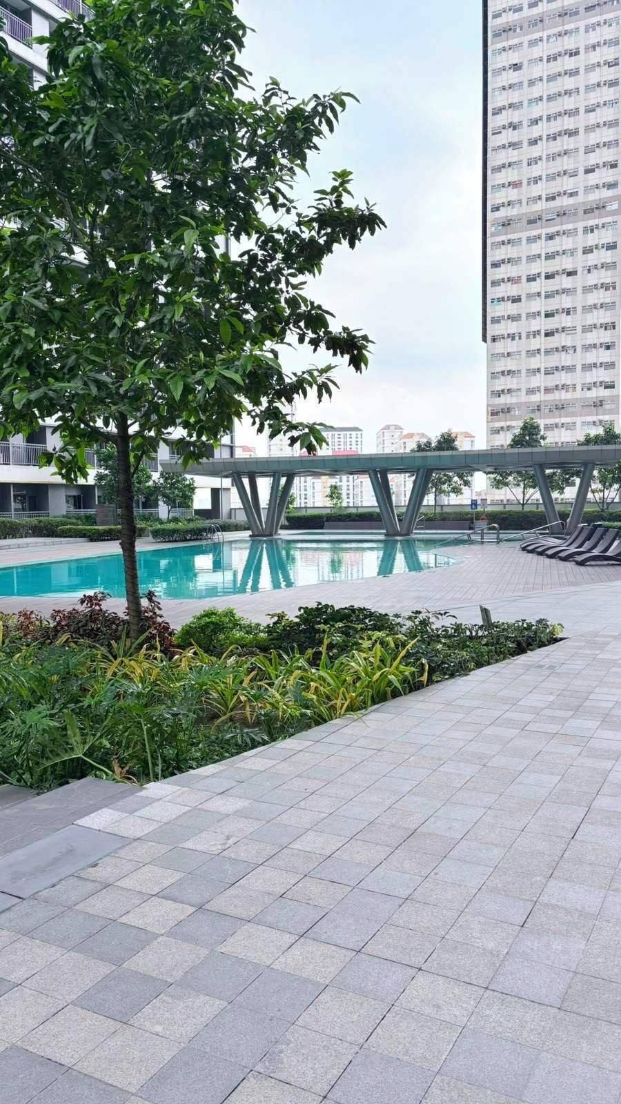 A modern outdoor pool area with a bridge, surrounded by greenery and a tall building in the background.