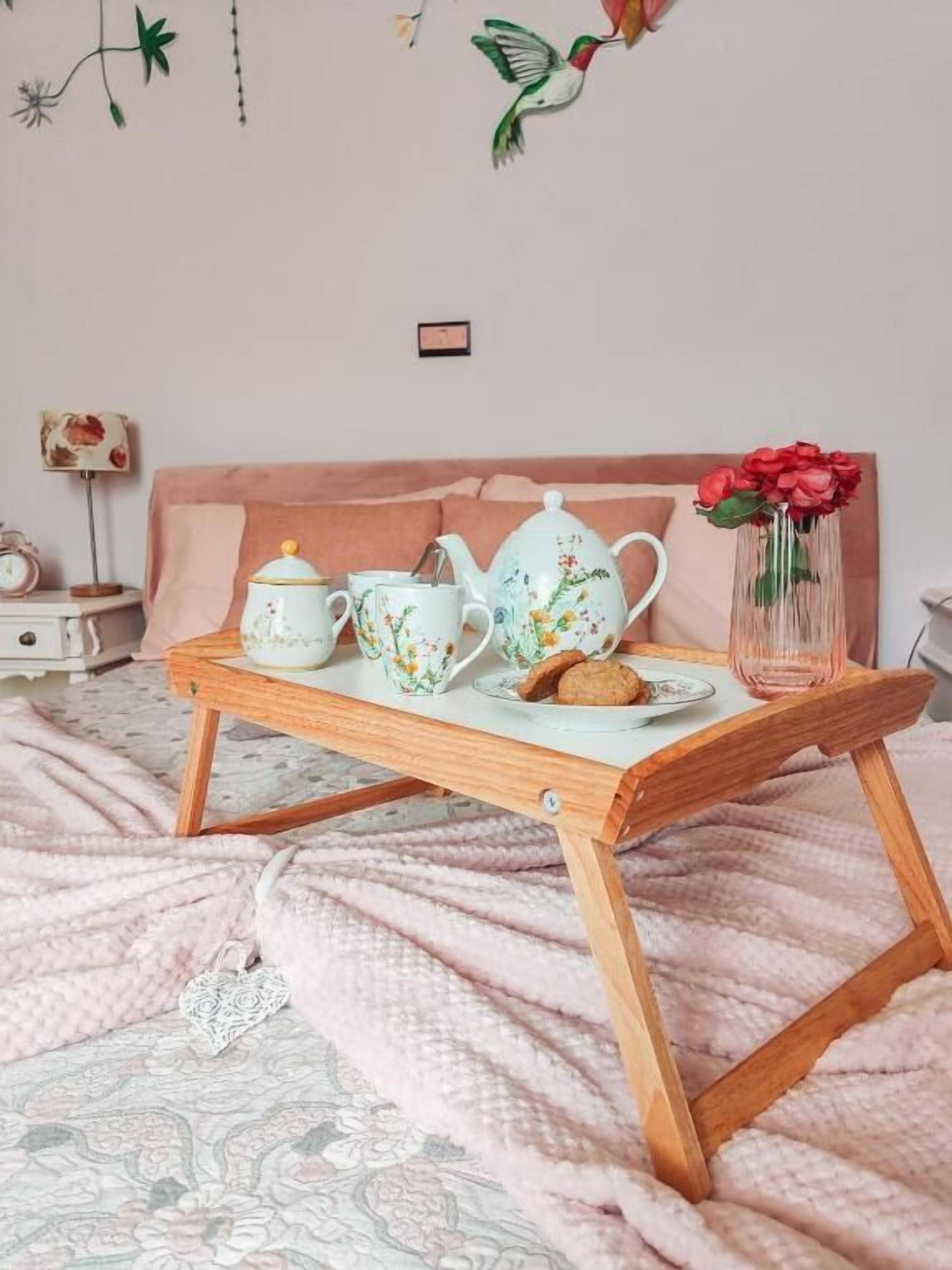 Una cama con un edredón rosa, una mesita de noche de madera con una bandeja que contiene una tetera, tazas y un florero con flores.
