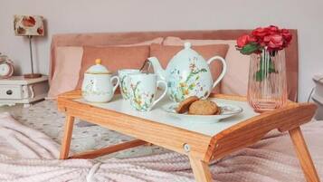 Una cama con un edredón rosa, una mesita de noche de madera con una bandeja que contiene una tetera, tazas y un florero con flores.