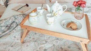 Una cama bien tendida con un cabecero rosa, una mesita de noche con una lámpara y una bandeja con juego de té y galletas.