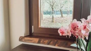 Un piso de madera, una ventana con vista a árboles y una planta en maceta con flores rosadas.