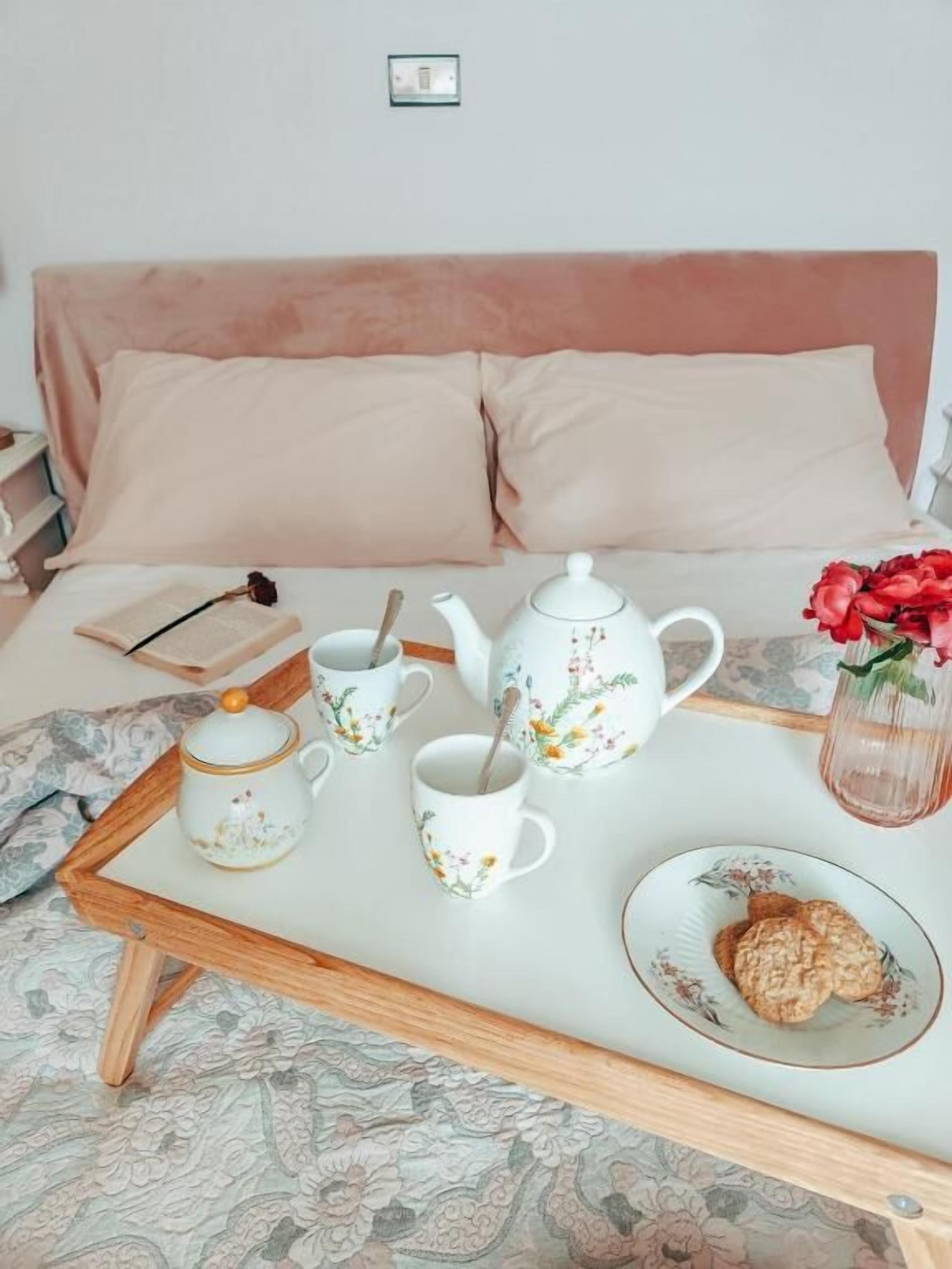 Una cama bien hecha con un cabecero rosa, una bandeja con tetera, galletas y un jarrón con flores.