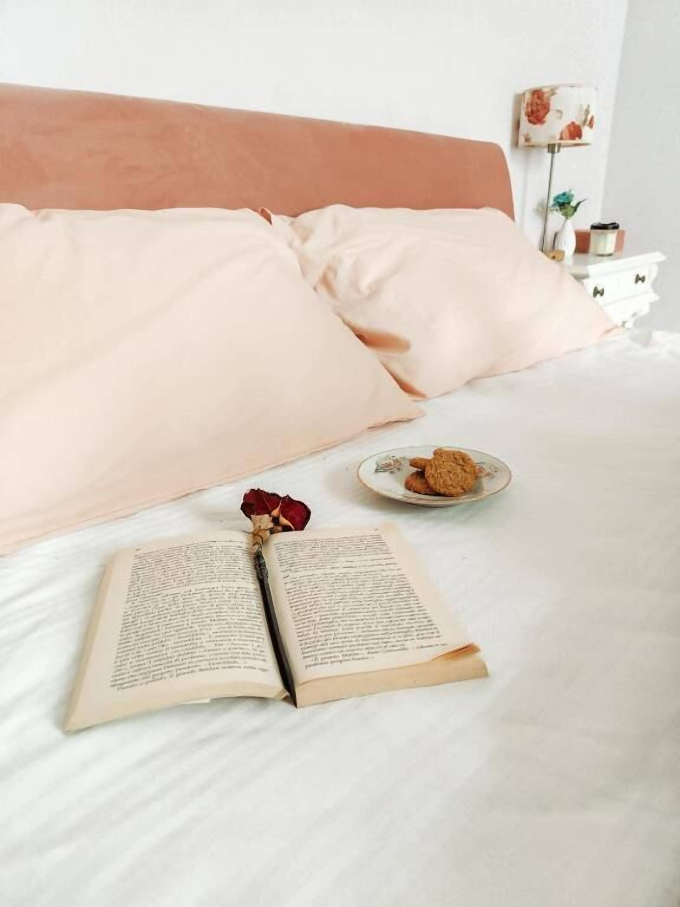 A bed with a book and a plate of cookies, a rose, and a lamp on a bedside table.