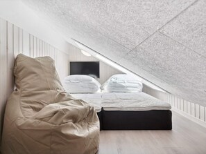 Ein Schlafzimmer mit schräg steilender Decke, einem Bett mit weißer Bettwäsche, einem schwarzen Ottomanen und einem beigen Sofa.