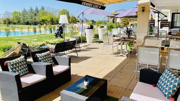Eine Terrasse im Freien mit Korbmöbeln, im Hintergrund ein Pool und ein Schild mit der Aufschrift 'VENDÉE SERIE'.