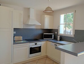Een kleine keuken met witte kastjes, een magnetron, een broodrooster, een waterkoker en een raam met uitzicht op groen.