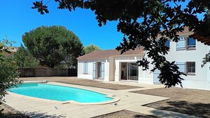 Ein Garten mit Swimmingpool, ein weißes Haus mit blauen Fensterläden und ein großer Baum.