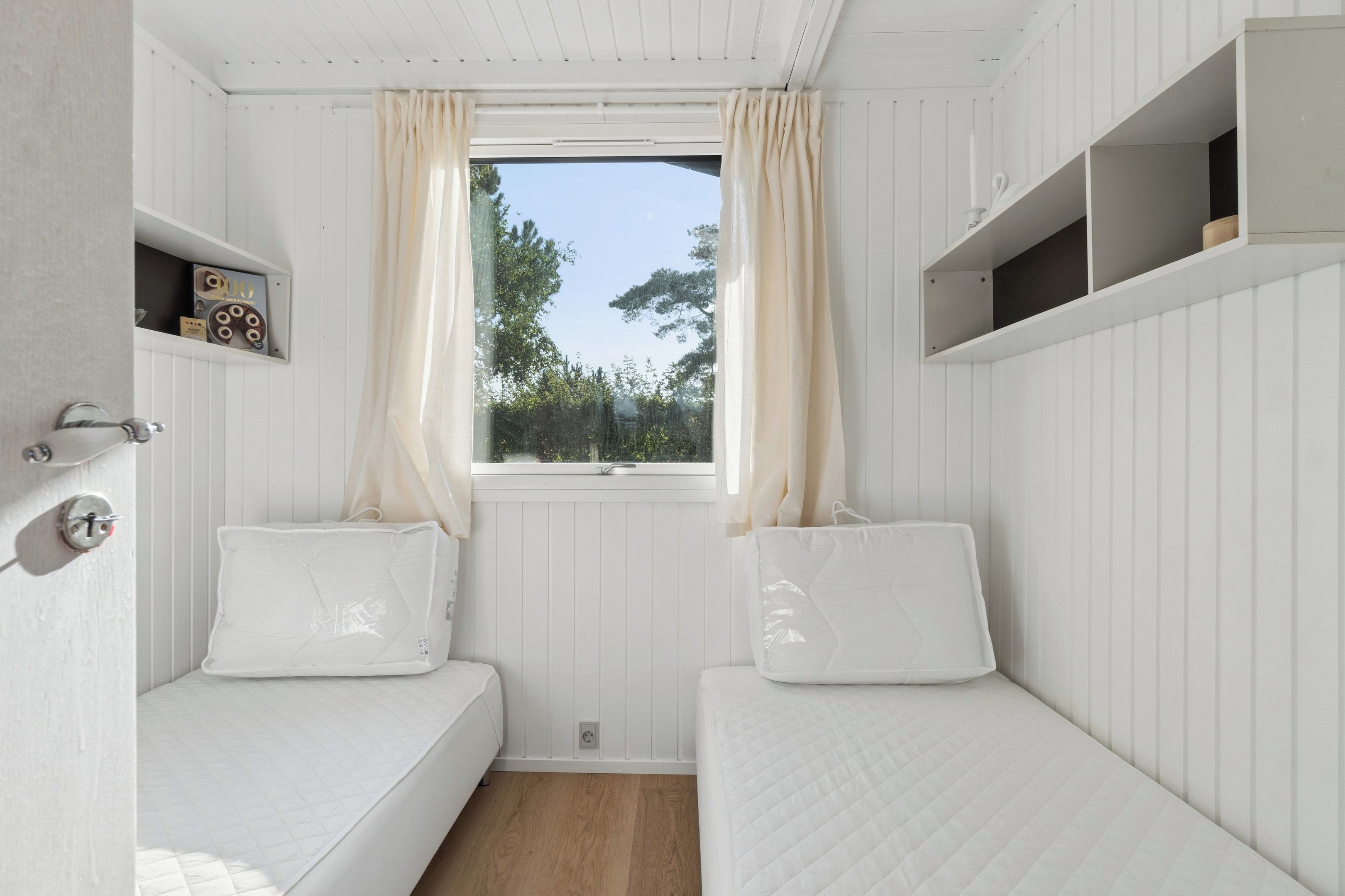 Une petite chambre à coucher décorée dans des tons de blanc, avec deux lits simples, une fenêtre avec des rideaux et une étagère ornée d’un objet décoratif.