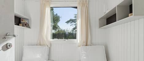 A small, white-themed bedroom with two single beds, a window with curtains, and a shelf with a decorative item.