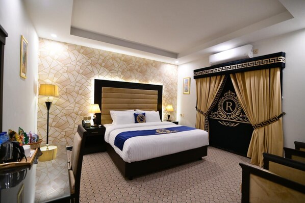 A hotel room with a large bed, a headboard, a nightstand, a desk with a lamp, and a decorative curtain with a monogram. - Royal Regency Hotel (Islamabad)