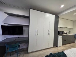 Een moderne kamer met een flatscreen televisie, een bureau, een blauwgroene fauteuil, een kledingkast en een kitchenette met een magnetron en een kleine koelkast.