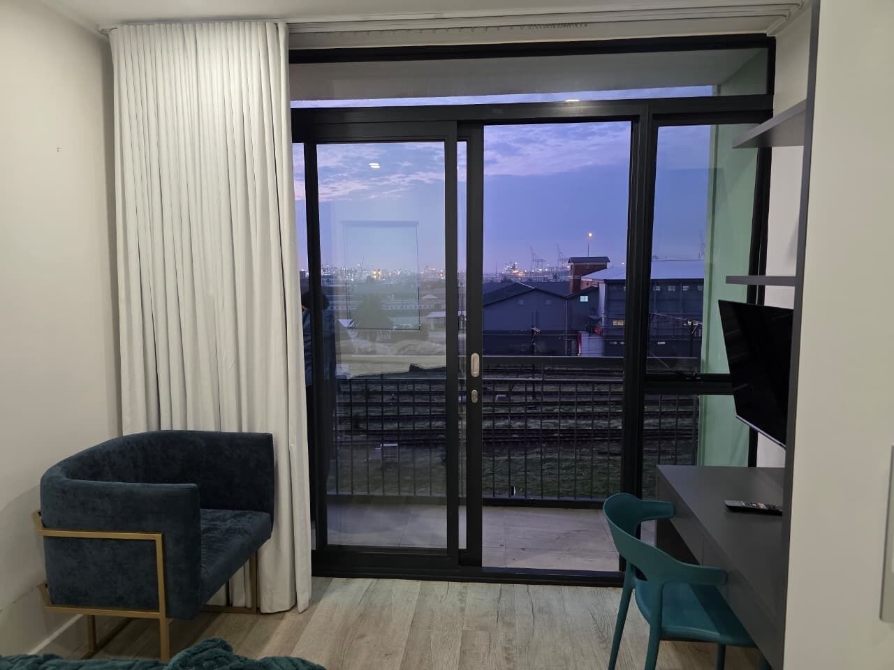 Una habitación con puerta de vidrio corredera que da a un balcón, un sillón azul oscuro, una silla verde azulada y un escritorio con televisor de pantalla plana.