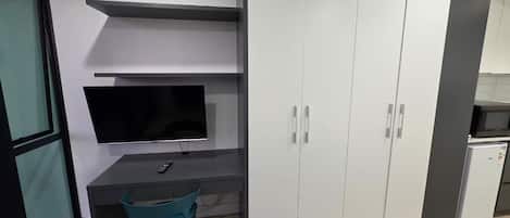 A compact room with a desk, a television, a wardrobe, and a small refrigerator.
