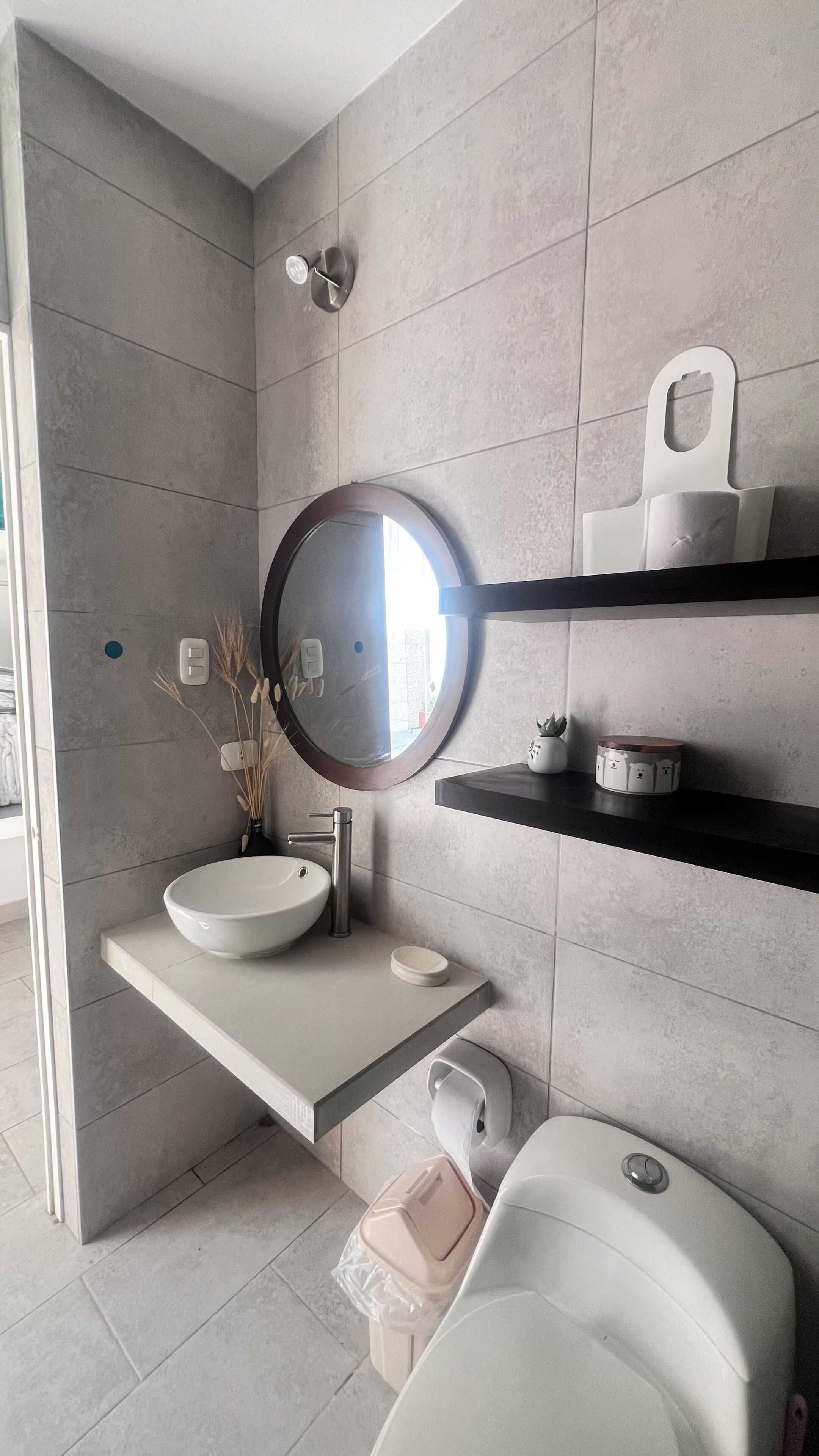 Et badeværelse med et rundt spejl, en vask, et toilet og en hylde med en beholder.