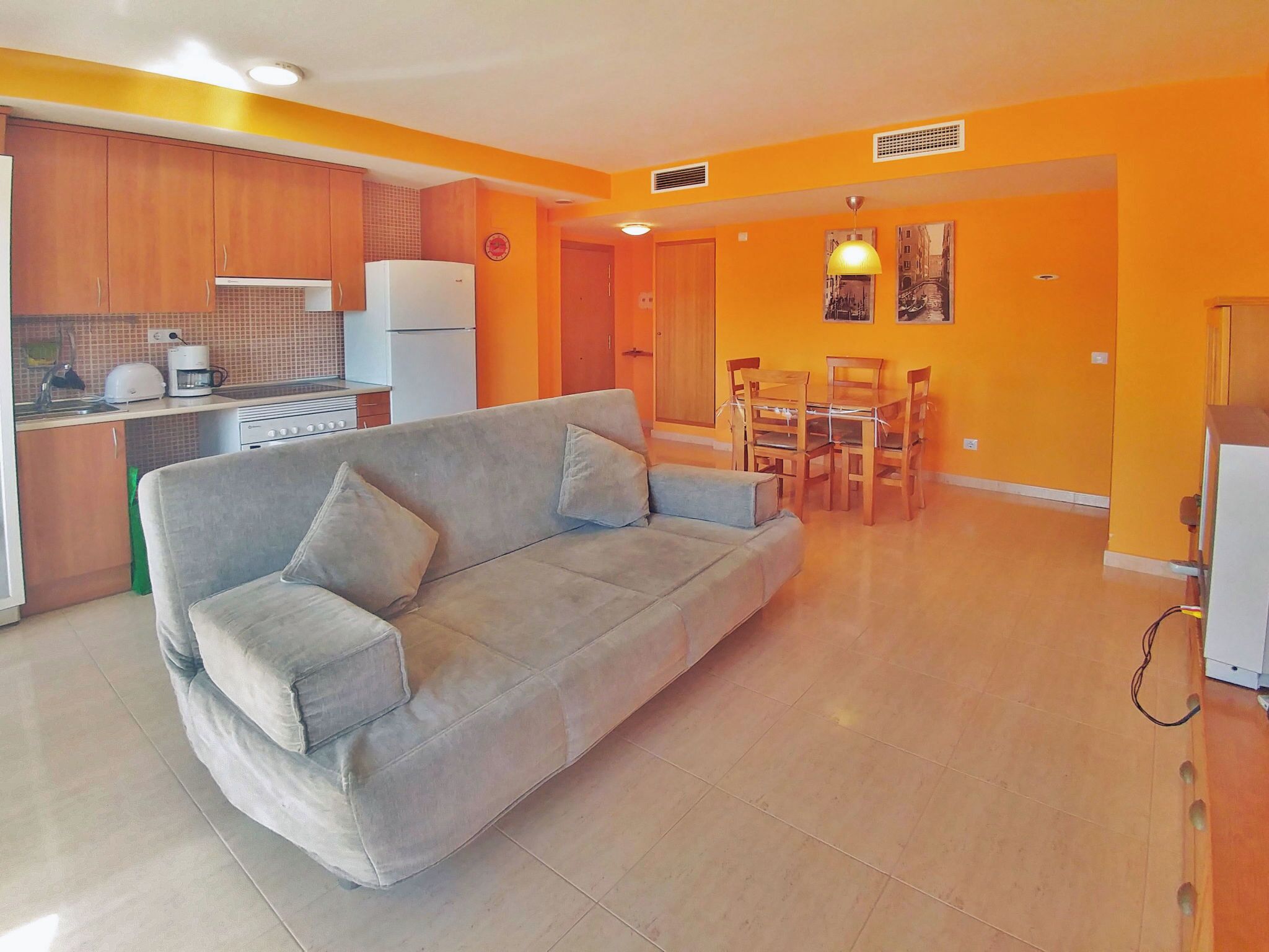 Ein Wohnzimmer mit Sofa, einem Esstisch mit Stühlen, einer Küchenzeile mit Schränken und Geräten sowie einem Flur mit einer Tür.