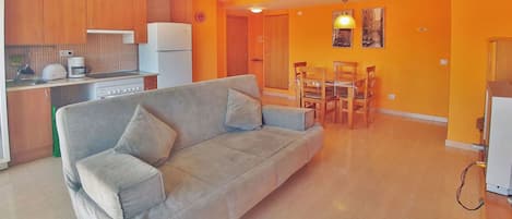 A living room with a sofa, a dining table with chairs, a kitchen area with cabinets and appliances, and a hallway with a door.