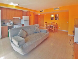 A living room with a sofa, a dining table with chairs, a kitchen area with cabinets and appliances, and a hallway with a door. - Coral I by Interhome (Miami Platja)