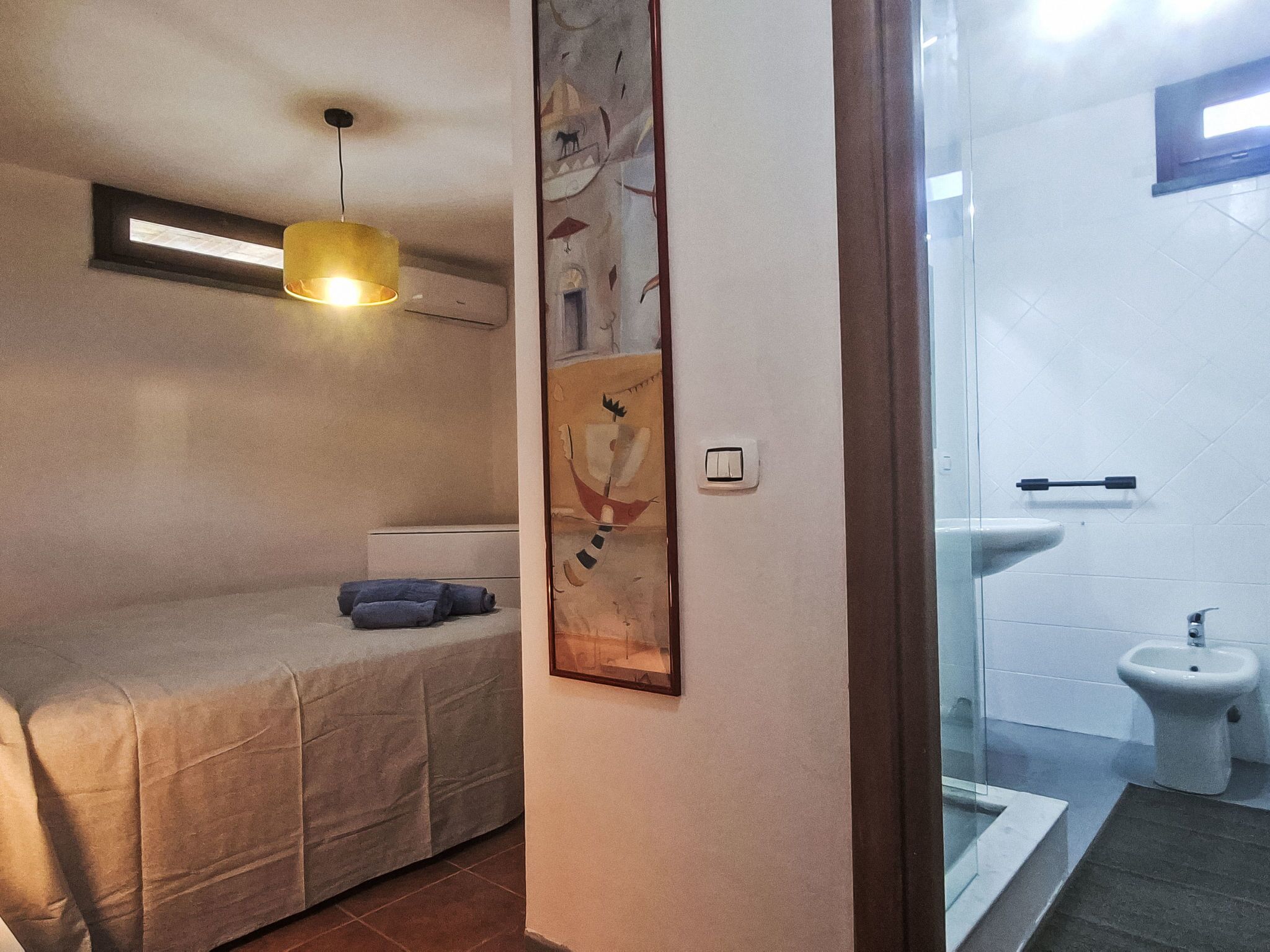 A bedroom with a bed, a bedside lamp, a wall-mounted air conditioner, and a window.