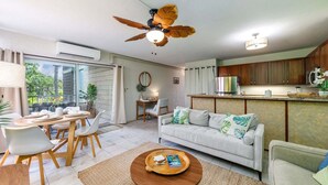 A modern living room with a sofa, dining table, and chairs, a ceiling fan, and a kitchen area with wooden cabinets. - Little Kona Getaway with AC & heated oceanfront pool 1 BD 1 BA with sleeper sofa (Kailua Kona)