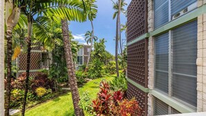 A lush garden with palm trees and tropical plants, a building with a grid-like facade, and a clear blue sky. - Little Kona Getaway with AC & heated oceanfront pool 1 BD 1 BA with sleeper sofa (Kailua Kona)