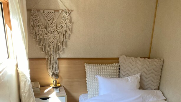 Un dormitorio con una cama, una mesita de noche con lámpara, un colgador de macramé y un cabecero de madera.