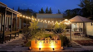 Un espacio exterior bien iluminado con un letrero que dice 'R GARDEN', rodeado por una terraza de madera y un sendero que lleva a un área de estar cubierta.