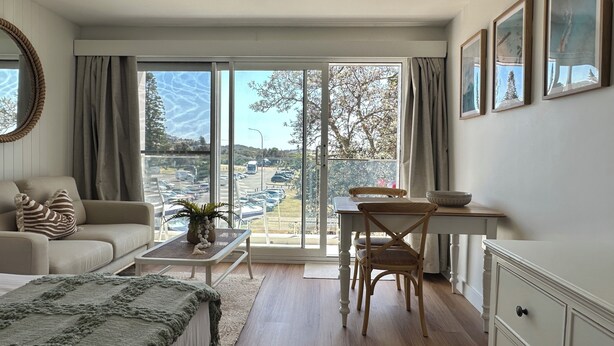 A room with a sofa, a dining table with chairs, a large window with a view of a parking lot and trees, and a cabinet with drawers. - Beachfront Studio, Views, Best Location (Dee Why)