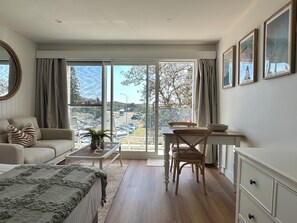 Una habitació amb un sofà, una taula de menjador amb cadires, una finestra gran amb vista a un aparcament i arbres, i un moble amb calaixos.