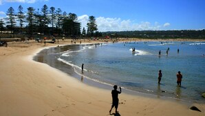 A beach with people walking along the shore and in the water, with trees and buildings in the background. - Beachfront Studio, Views, Best Location (Dee Why)