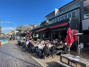 An outdoor dining area with tables and chairs, a wooden deck, and a building with a red awning. - Beachfront Studio, Views, Best Location (Dee Why)